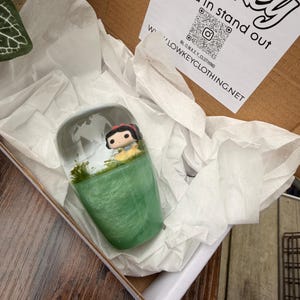 May include: A clear, cylindrical resin art piece featuring a Snow White Funko Pop figure set against a green, glittery base. The piece is packaged in a cardboard box with white tissue paper and a printed label that reads "Lowkey".