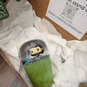 May include: A clear, oval-shaped decorative item with a small figurine on a green base. The figurine is a cartoon character. The item is in a cardboard box with white tissue paper.