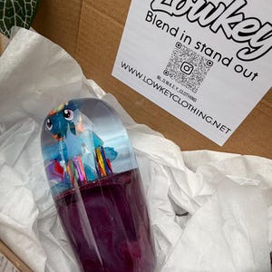 May include: A clear, capsule-shaped resin art piece with a blue and purple interior, featuring a colorful cartoon character. The capsule is set in a box with white tissue paper and a label that reads "Lowkey Blend in stand out".
