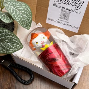 以下が含まれることがあります： 赤と透明のタンブラーの中にキャラクターフィギュアが入ったノベルティグッズ。白い箱にティッシュペーパーを入れてあります。箱には、黒い編み込みキーホルダーと「Lowkey Blend in stand out」と書かれたステッカーも入っています。