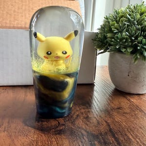 May include: A clear, cylindrical resin gear shift knob featuring a yellow Pikachu character figurine embedded inside. The base of the knob displays a swirling pattern of dark blue, black, and yellow colors, creating a deep, marbled effect.
