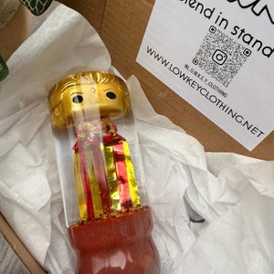 May include: A Funko Pop figure with a gold head and a red and gold body is encased in a clear cylinder. The figure sits on a red base. The packaging includes white paper and a cardboard box. The text "Blend in stand" and a website address are visible.