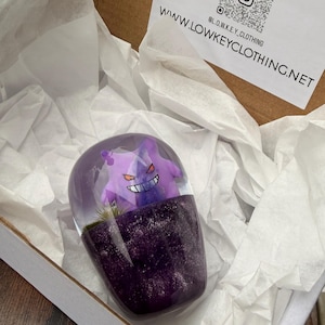 May include: A purple capsule-shaped resin art piece featuring a Gengar figure inside. The base is a darker purple with glitter, and the top is clear. The piece is presented in a white box with tissue paper. The website address is visible.