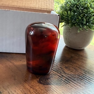 May include: A smooth, rounded gear shift knob with a deep, translucent amber and dark brown marbled pattern. The object has a glossy, reflective surface and a tapered cylindrical shape, showcasing a rich, multi-tonal resin appearance.