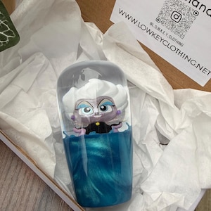 Puede incluir: Un vaso transparente con un diseño de personaje de dibujos animados. El personaje tiene cabello blanco, piel morada y ojos azules. La parte inferior del vaso está llena de un líquido azul arremolinado. El vaso está dentro de una caja de cartón con papel de seda blanco.