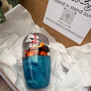 Op de afbeelding: Een heldere harsbeker met een wit en rood gehelmde figuur, een oranje en bruin landschap en een glinsterende blauwe basis. De beker wordt gepresenteerd in een wit gevoerde doos. De tekst op de doos luidt "Blend in stand out".