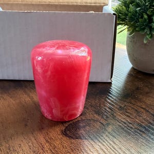May include: A cylindrical gear shift knob featuring a vibrant hot pink and white marbled resin finish. The surface is smooth and glossy, displaying a swirling pattern of opaque pink and translucent white tones throughout the solid material.