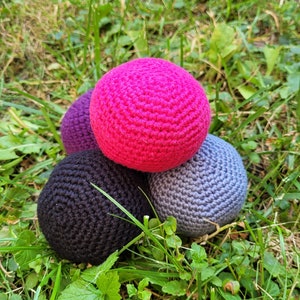 Puede incluir: Cuatro bolas de crochet en diferentes colores: rosa, morado, negro y gris. Las bolas están dispuestas en forma de pirámide sobre un lecho de hierba verde.