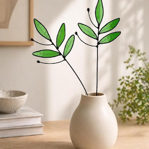 May include: A small, round, off-white ceramic vase containing two decorative stems. Each stem features thin black metal wire branches with multiple translucent green glass leaves and small black spherical accents at the tips of the branches.