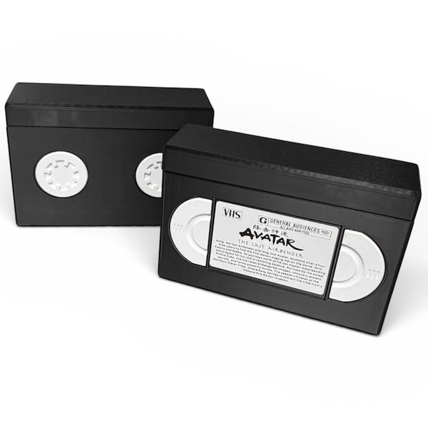 May include: Two black VHS tape-shaped boxes. One box is angled towards the viewer, displaying a white label with the text "AVATAR THE LAST AIRBENDER". The other box is in the background, showing the circular tape reels.