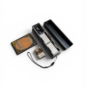 Puede incluir: Una caja negra para guardar cartas con una tapa abierta, que contiene una baraja de cartas, dados y una carta. Una carta de Magic: The Gathering está al lado de la caja, junto con una pequeña baraja de cartas. Una correa negra está unida a la caja.