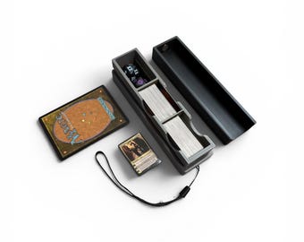 Könnte beinhalten: Eine schwarze Kartenaufbewahrungsbox mit offenem Deckel, die ein Kartenspiel, Würfel und eine Karte enthält. Eine Magic: The Gathering-Karte befindet sich neben der Box, zusammen mit einem kleinen Kartenspiel. Ein schwarzes Schlüsselband ist an der Box befestigt.