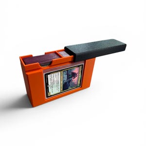 Peut inclure: Une boîte de rangement pour cartes à collectionner en plastique orange avec un couvercle noir coulissant. La boîte présente une découpe frontale rectangulaire affichant une carte avec le texte "Affinity for artifacts" et "Flying, deathtouch".