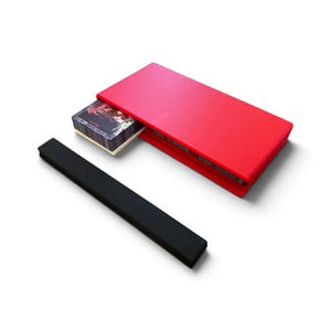 May include: A rectangular red storage box with a sliding lid, partially open to reveal a stack of rectangular playing cards with dark illustrated backs. A separate, long, narrow black rectangular bar is positioned next to the red box.