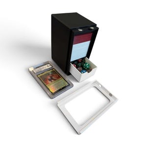 K&ouml;nnte beinhalten: Eine schwarze, vertikale Kartenaufbewahrungsbox mit einer wei&szlig;en Schublade, die verschiedene bunte Spielw&uuml;rfel enth&auml;lt. Eine bewertete Sammelkarte in einer transparenten Kunststoffh&uuml;lle und ein separater wei&szlig;er Rahmen liegen daneben.
