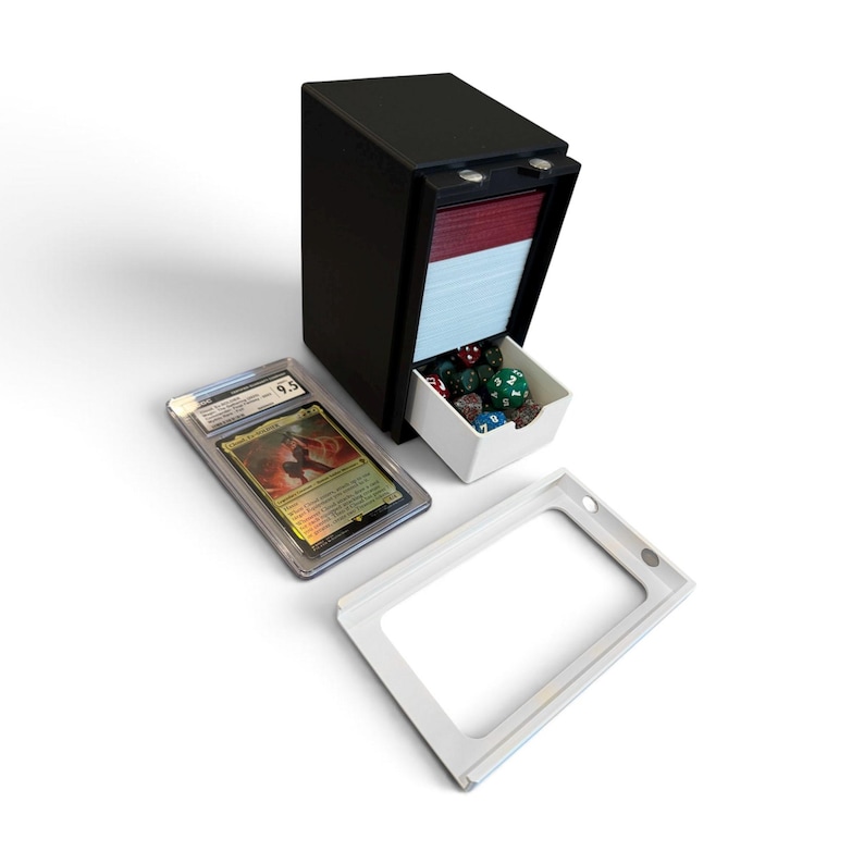 K&ouml;nnte beinhalten: Eine schwarze, vertikale Kartenaufbewahrungsbox mit einer wei&szlig;en Schublade, die verschiedene bunte Spielw&uuml;rfel enth&auml;lt. Eine bewertete Sammelkarte in einer transparenten Kunststoffh&uuml;lle und ein separater wei&szlig;er Rahmen liegen daneben.