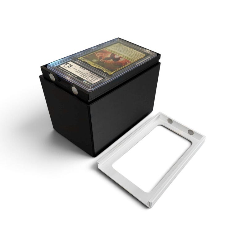 K&ouml;nnte beinhalten: Eine schwarze, rechteckige Aufbewahrungsbox mit magnetischem Deckel, die eine einzelne bewertete Sammelkarte in einer transparenten Kunststoffh&uuml;lle enth&auml;lt. Die H&uuml;lle tr&auml;gt ein CGC-Etikett mit der Note 9,5. Ein separater wei&szlig;er Rahmen liegt daneben.