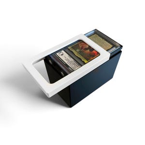 May include: A black rectangular storage box with a white sliding lid featuring a rectangular cutout. Inside the box, two graded trading cards in clear protective plastic cases are visible, with the top card showing a 9.5 grade and text.