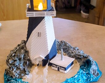 May include: A decorative lighthouse model with a blue and white color scheme. The lighthouse has a light inside, and sits on a base with a small building, rocks, and a boat. The base is surrounded by blue water.