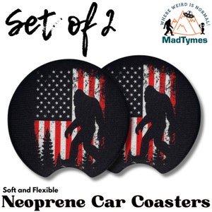 May include: Set of two black neoprene car coasters with a red, white, and blue American flag design and a silhouette of Bigfoot walking in front of the flag. The text "Where Weird Is Normal!" and "MadTymes" are printed on the coasters. The coasters are soft and flexible.