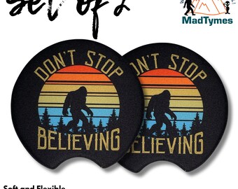 May include: Set of two black round neoprene car coasters with a retro sunset design. The coasters feature a silhouette of Bigfoot walking through a forest with the text "Don't Stop Believing".