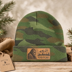 May include: A green, brown, and black camouflage knit beanie featuring a rectangular tan leather patch on the cuff. The patch displays an engraved graphic of a Bigfoot, mountains, a moon, and the text "MINDSPEAK" with a small pine tree icon.