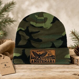 May include: A camouflage beanie with a green, black, and brown pattern. The cuff features a rectangular leather patch with a black graphic of a winged creature, a bridge, and the text "NIGHT SHIFT MOTHMAN" printed in a dark font.
