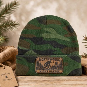 May include: A green, black, and brown camouflage knit beanie with a folded cuff. A rectangular brown leather patch is centered on the cuff, featuring a black graphic of a creature, trees, and a tower with the text NORTHWOODS NIGHT PATROL.
