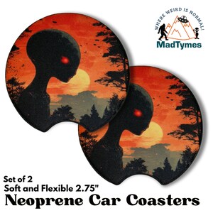 May include: Set of two neoprene car coasters with a sunset alien design. The coasters are soft and flexible, measuring 2.75 inches. The design features a black alien silhouette with red eyes against an orange and yellow sunset.