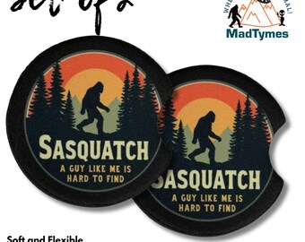 May include: Set of two black neoprene car coasters. Each coaster features a Sasquatch silhouette walking in front of a sunset and forest scene. Text reads "Sasquatch, A guy like me is hard to find."