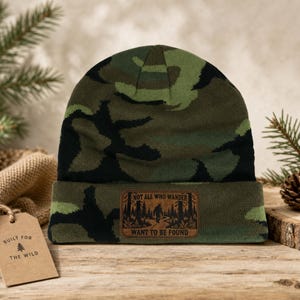 May include: A camouflage beanie in shades of green, black, and brown. The cuff features a rectangular brown patch with a black graphic of a forest and a creature, with the text "NOT ALL WHO WANDER WANT TO BE FOUND" printed on the patch.