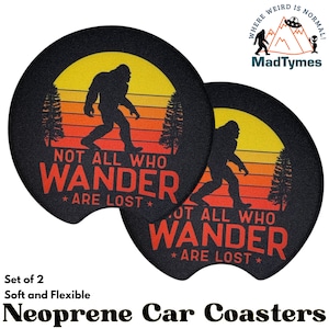 May include: Set of two black round neoprene car coasters with a sunset graphic, a silhouette of Bigfoot, and the text "Not all who wander are lost".