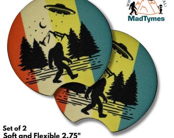 May include: Set of two 2.75 inch round neoprene car coasters with a retro design. The coasters feature a yellow, orange, and blue striped background with a silhouette of Bigfoot walking through a forest with a UFO flying overhead. The text "Where Weird Is Normal! MadTymes" is printed on the coasters.