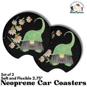 May include: Set of two 2.75 inch round black neoprene car coasters with a gnome and flowers design. The gnome is wearing a green hat and has a white beard. The flowers are white with yellow centers. The background is a black galaxy print. The text "Where Weird is Normal" and "MadTymes" is printed on the top of the image.