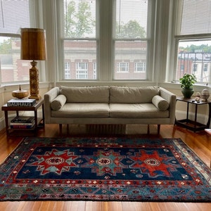 May include: A white sofa with a blue and red patterned rug in front of it. The sofa has a wooden frame and a light brown lamp with a gold base is next to it.