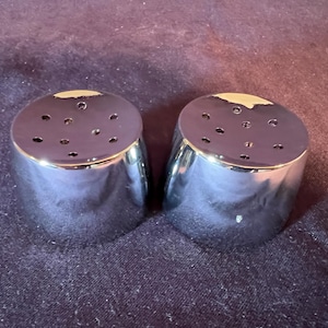 May include: A pair of cylindrical salt and pepper shakers with a polished chrome finish. Each shaker features a flat top with a circular pattern of small dispensing holes. The metallic surface is reflective, showing light highlights on the top edges.