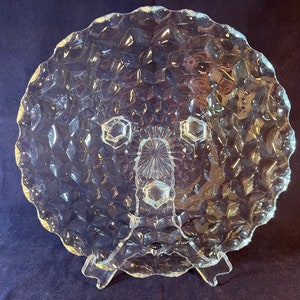May include: A clear glass serving dish with a diamond pattern and three feet. The dish is made of clear glass and has a scalloped edge.