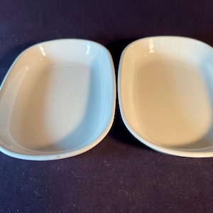 May include: Two white, oval-shaped serving dishes with rounded corners. The dishes are empty and have a smooth, glossy finish. They are placed side by side on a dark blue surface. The dishes are likely made of ceramic or porcelain.