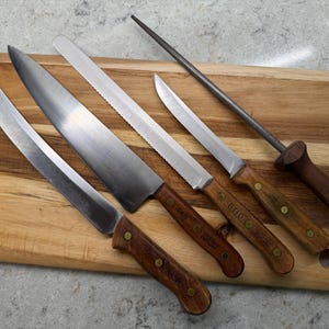 May include: A collection of kitchen knives and a sharpening steel rest on a wooden cutting board. The knives have stainless steel blades and wooden handles. The cutting board is rectangular with rounded corners.
