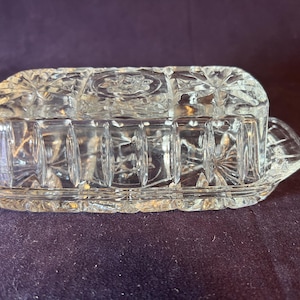 May include: A clear, rectangular glass butter dish with a lid. The dish and lid feature a starburst and geometric pattern. The dish has a tray base with handles on each end. The butter dish is sitting on a dark purple surface.