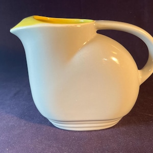 May include: A white ceramic pitcher with a curved handle and a yellow rim. The pitcher has a rounded body and a spout for pouring. The design is simple and elegant, suitable for serving beverages.
