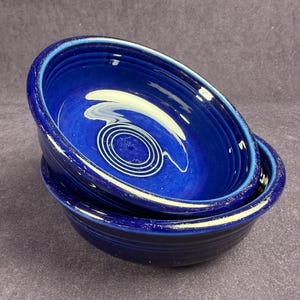May include: Two cobalt blue ceramic bowls are stacked. The bowls have a glossy finish and a white concentric circle design in the center. The rims of the bowls have a lighter blue color.