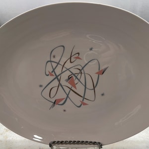 May include: Oval serving platter with a white background and a mid-century modern design. The design features abstract lines in brown and blue, with pink triangles and small star accents. A decorative piece for serving.