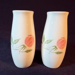 May include: Two white ceramic salt and pepper shakers. Each shaker features a pink rose design with green leaves and stems. The shakers have a slightly flared top and a smooth, glossy finish. They are likely vintage or retro.