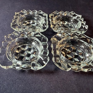 May include: Four clear glass candle holders with a diamond pattern in the center and a scalloped edge. The candle holders are arranged in a square formation on a dark blue surface.