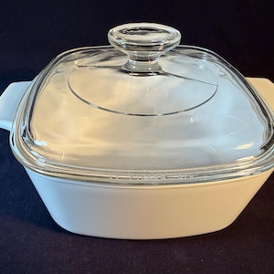 May include: Square, white casserole dish with a clear glass lid. The dish has two white handles and a clear glass knob on the lid. The dish is likely made of ceramic or glass and is designed for baking or serving food.