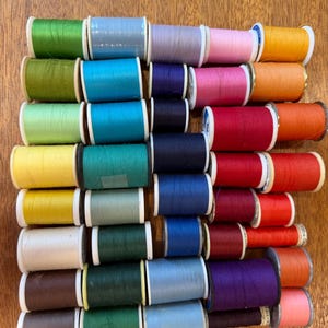May include: A collection of sewing thread spools arranged in a grid, featuring a wide variety of solid colors including shades of green, blue, red, yellow, pink, purple, orange, brown, and white, each wound on a standard plastic spool.