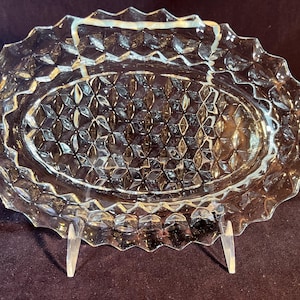 May include: Clear, oval-shaped glass serving dish with a diamond pattern in the center and a scalloped edge. The dish is displayed on a clear stand, showcasing its intricate design. A decorative piece for serving appetizers or desserts.