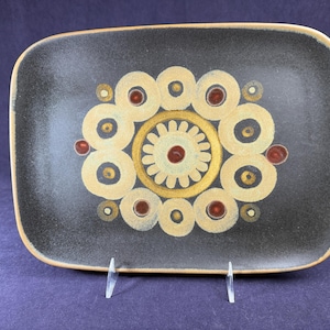 May include: Rectangular ceramic serving dish with a dark gray background and a beige and gold floral design. The dish has a slightly raised edge and is supported by a clear plastic stand. The floral design features concentric circles and dots.