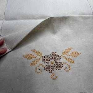 May include: Off-white linen table runner with cross-stitched floral designs in brown and orange. The runner features a folded section, showcasing the detailed embroidery and a decorative stitched border.